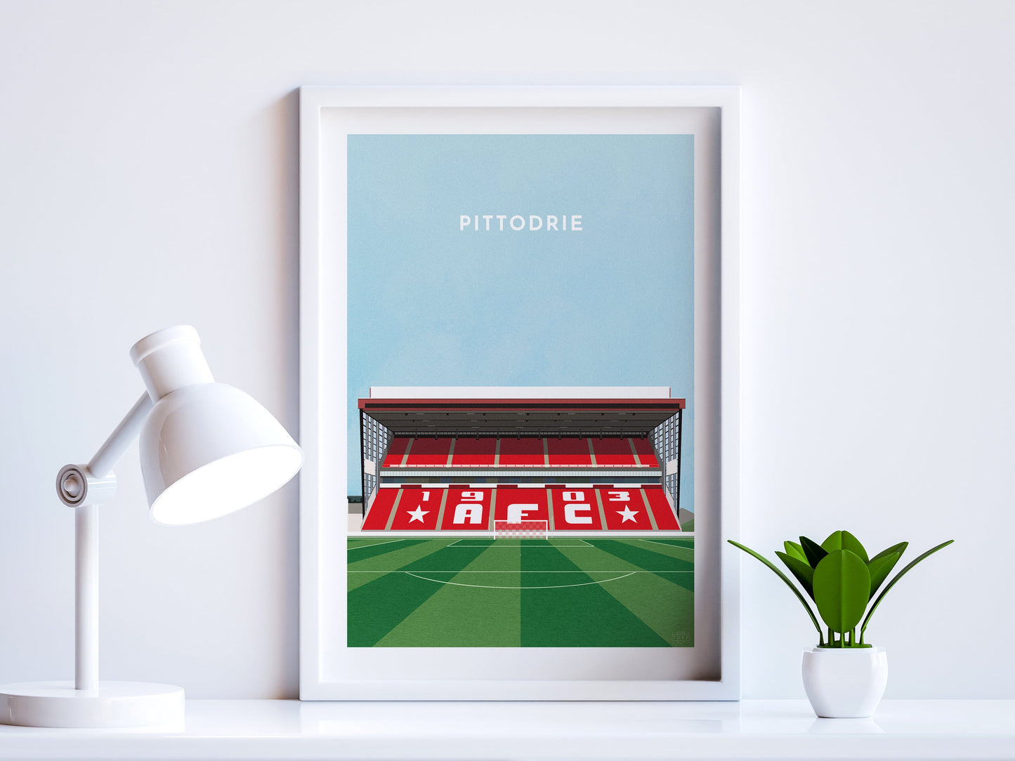 A contemporary style illustration of Aberdeen's football stadium, Pittodrie, displayed as an art print within a white frame in a minimalist home office setting.