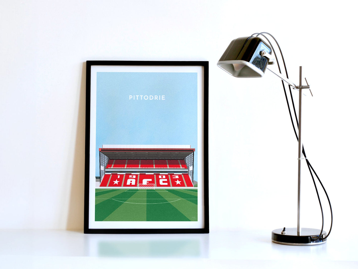 A contemporary style illustration of Aberdeen's football stadium, Pittodrie, displayed as an art print within a black frame in a minimalist home office setting.