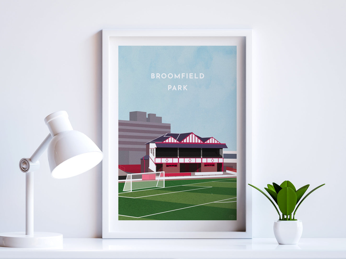 A contemporary style illustration of Airdrieonians' former football stadium, Broomfield Park, displayed as an art print within a white frame in a minimalist home office setting.