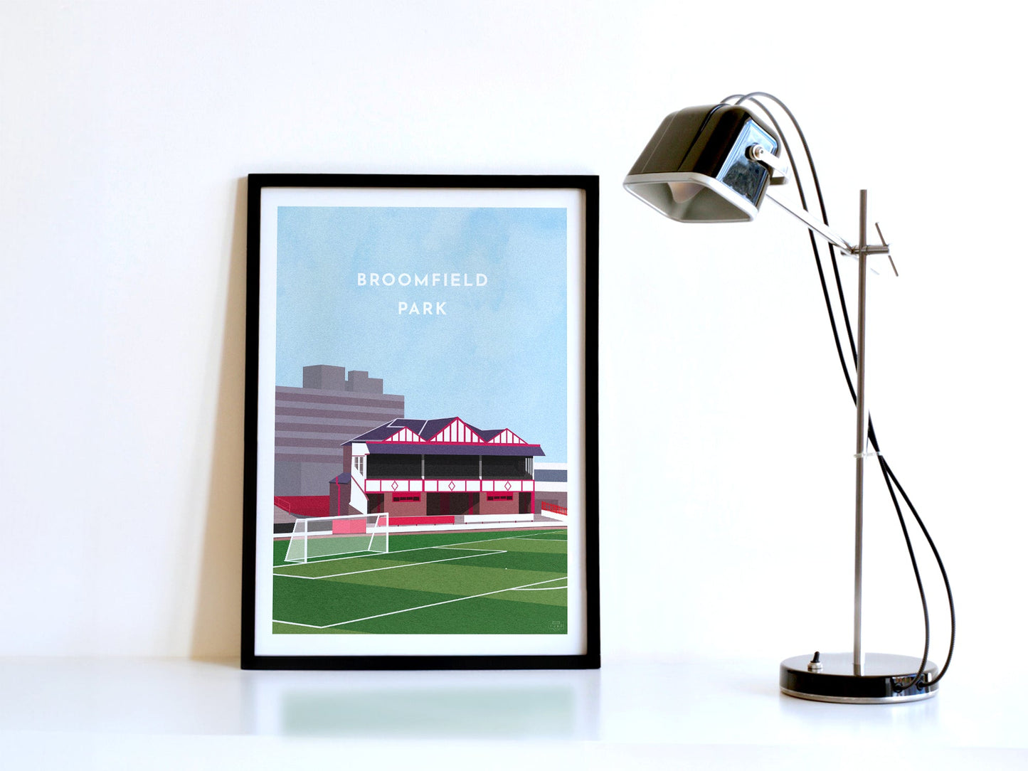 A contemporary style illustration of Airdrieonians' former football stadium, Broomfield Park, displayed as an art print within a black frame in a minimalist home office setting.
