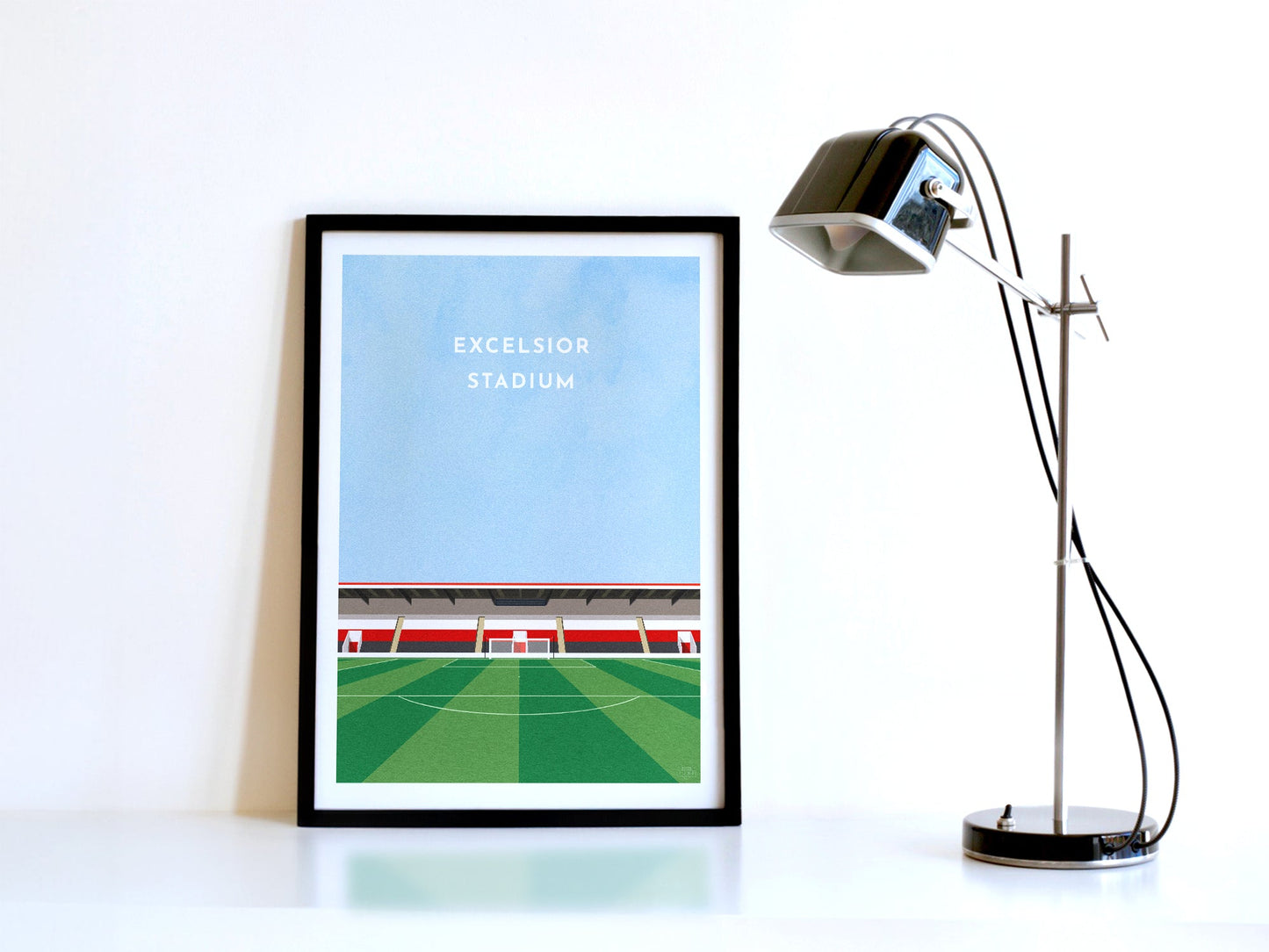 A contemporary style illustration of Airdrieonians' football stadium, the Excelsior Stadium, displayed as an art print within a black frame in a minimalist home office setting.