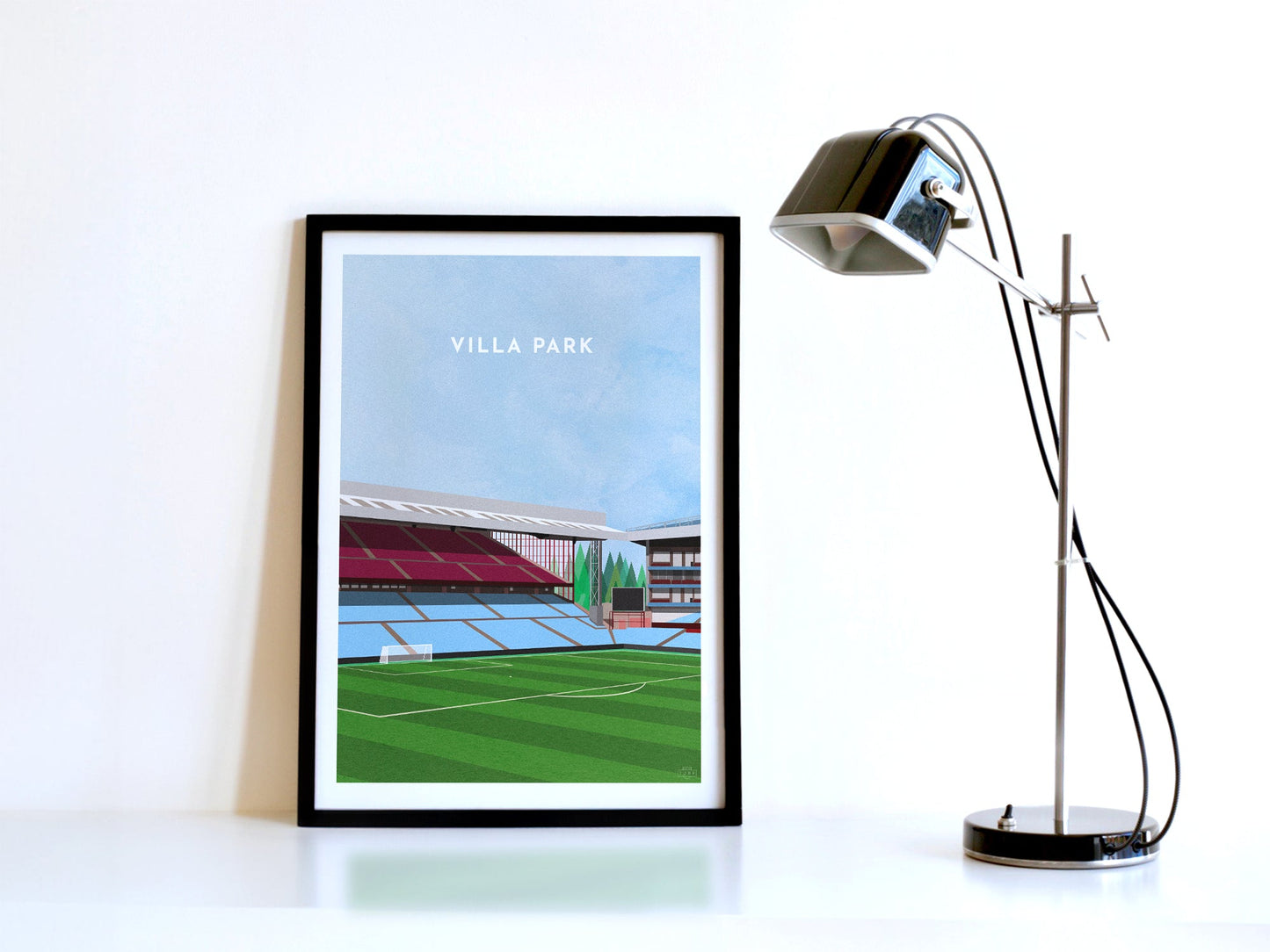 A contemporary style illustration of Aston Villa's football stadium, Villa Park, displayed as an art print within a black frame, next to a retro style black lamp.
