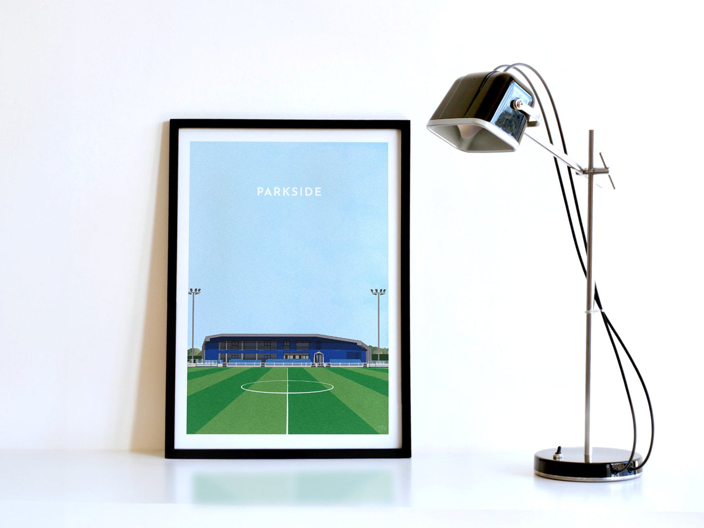 A contemporary style illustration of Aveley's football stadium, Parkside, displayed as an art print within a black frame in a minimalist home office setting.