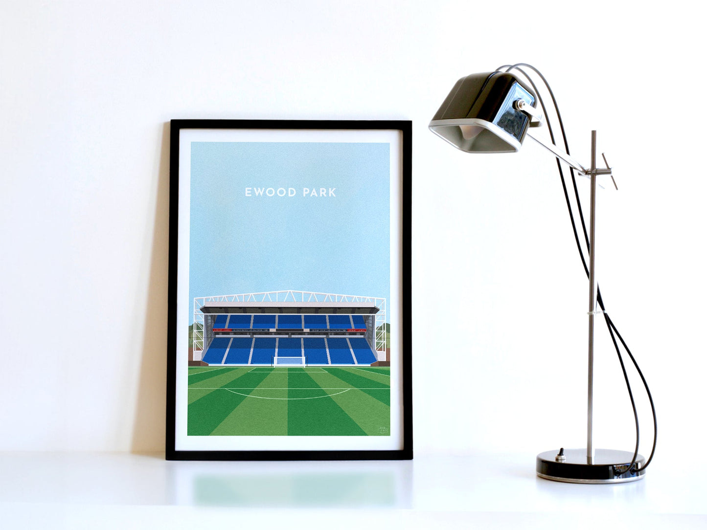A contemporary style illustration of Blackburn Rovers' football stadium, Ewood Park, displayed as an art print within a black frame in a minimalist home office setting.