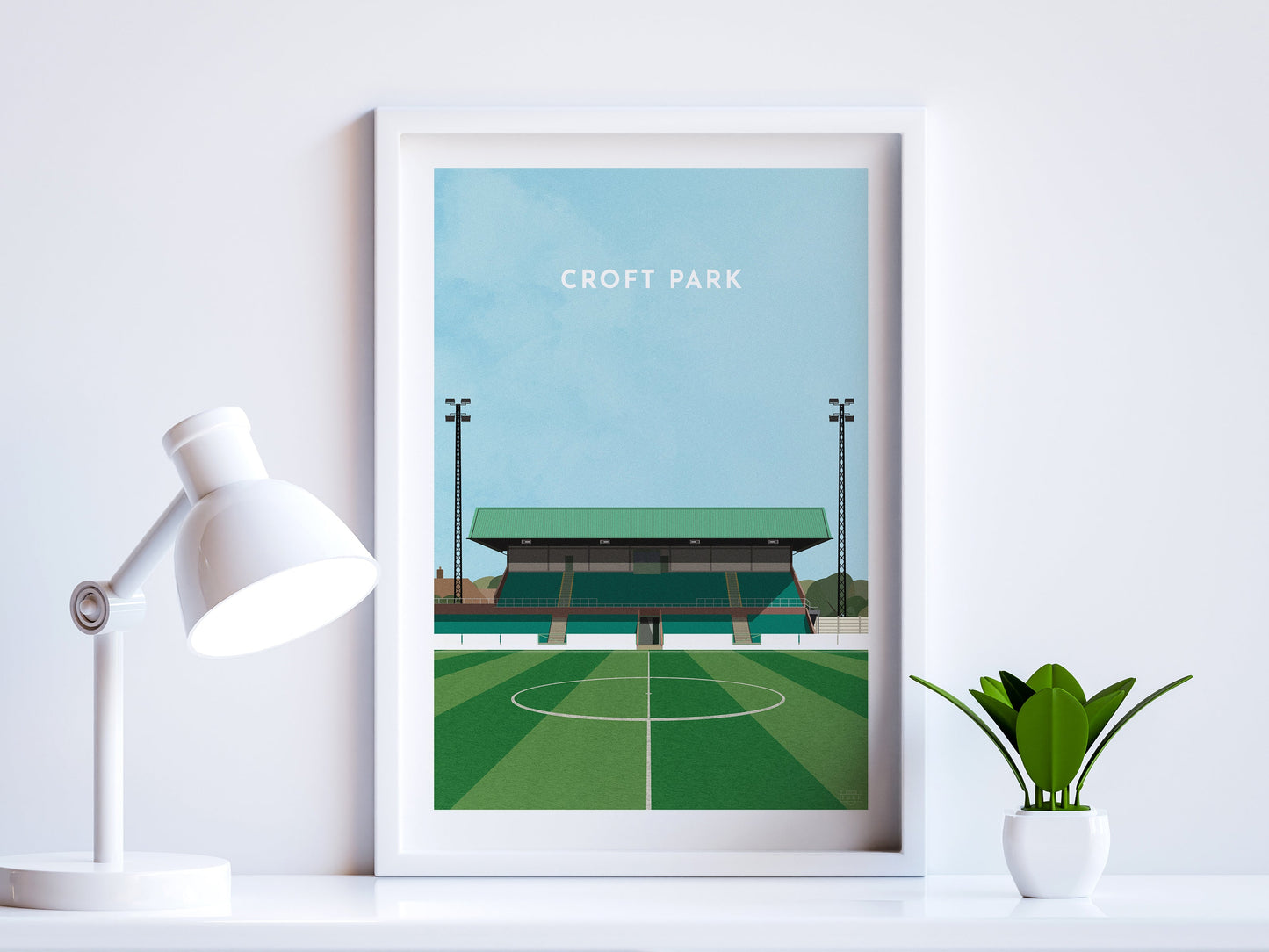 A contemporary style illustration of Blyth Spartans' football stadium, Croft Park, displayed as an art print within a white frame in a minimalist home office setting.