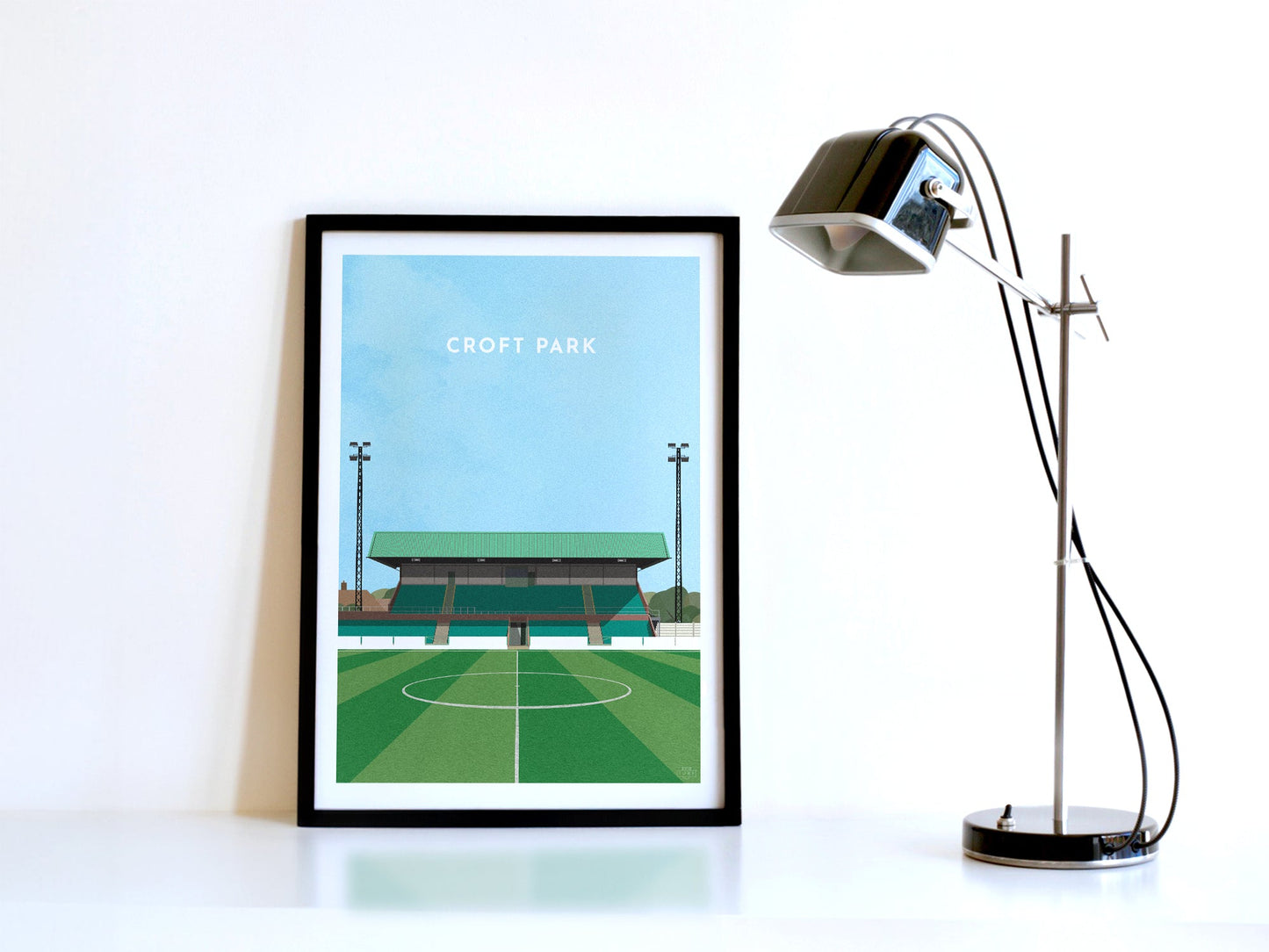 A contemporary style illustration of Blyth Spartans' football stadium, Croft Park, displayed as an art print within a black frame in a minimalist home office setting.