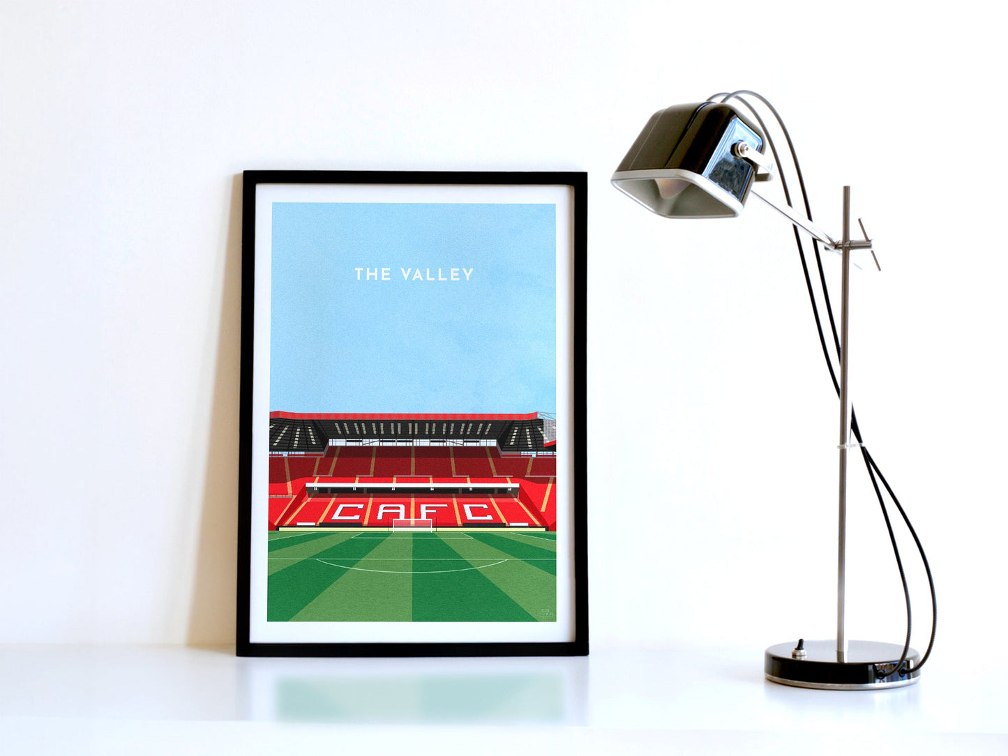 A contemporary style illustration of Charlton Athletic's football stadium, The Valley, displayed as an art print within a black frame in a minimalist home office setting.