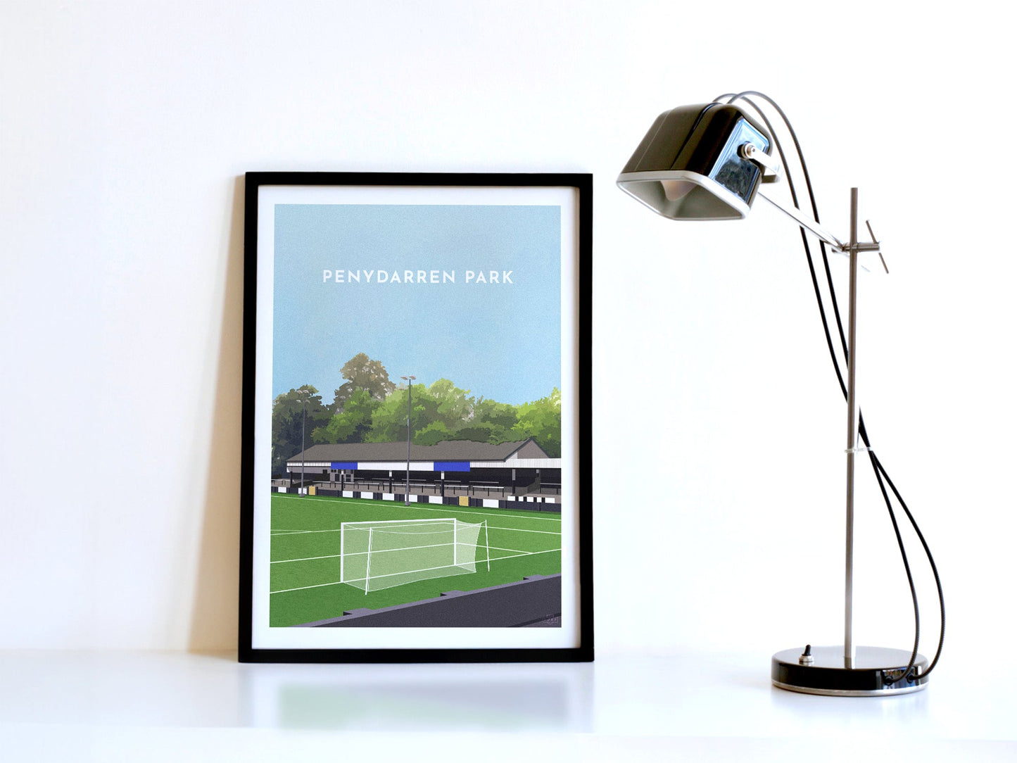 A contemporary style illustration of Merthyr Tydfil's football ground, Penydarren Park, displayed as an art print within a black frame, next to a retro style black lamp.