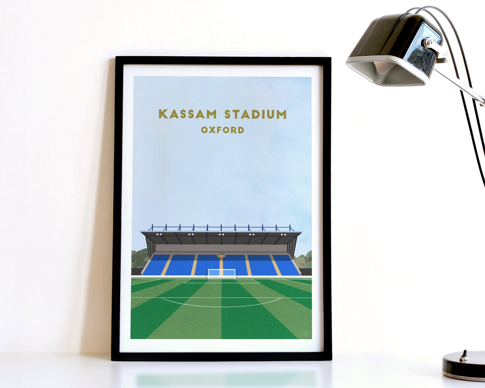 A modern style Oxford United Print, featuring a contemporary illustration of their home stadium the Kassam Stadium in Oxford, displayed within a black frame.