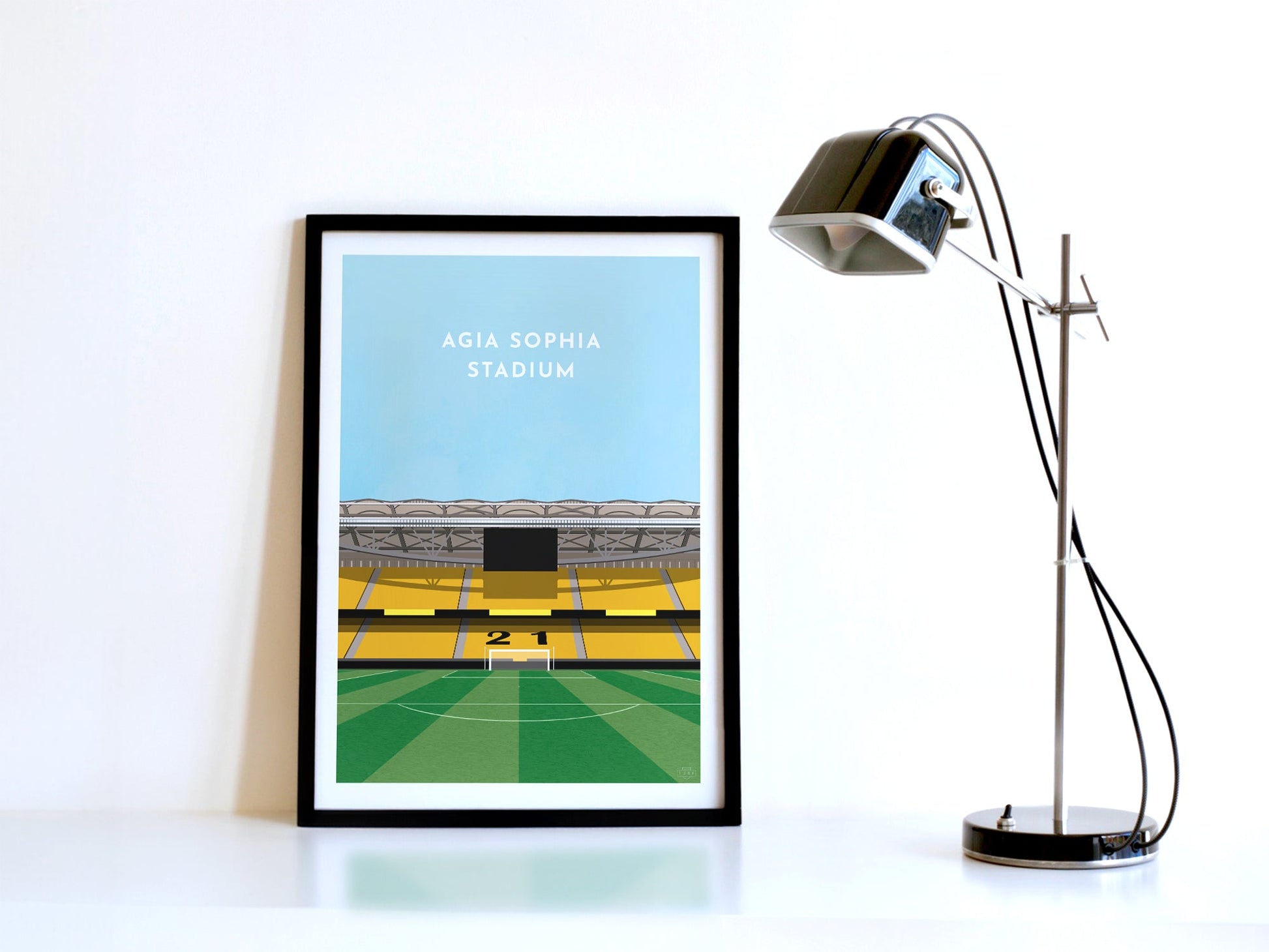 A contemporary style illustration of AEK Athens football stadium, the Agia Sophia Stadium, displayed as an art print within a black frame in a minimalist home office setting. 