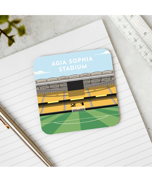 Square hardwood coaster placed on a desk, with glossy sublimation printed finish featuring contemporary illustration of Agia Sophia Stadium in Nea Filadelfia, home to AEK Athens, showcasing a minimalist design with vibrant yellow and black stands, green football pitch, under a blue sky.