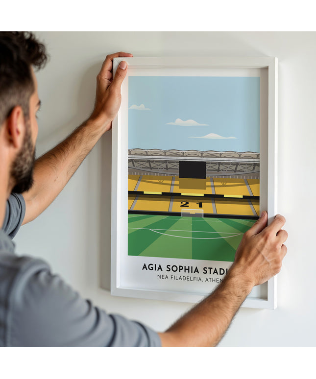 A man hanging a framed art print, featuring a contemporary style illustration of Agia Sophia Stadium, home to AEK Athens.