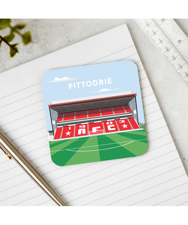 Square hardwood coaster placed on a desk, with glossy sublimation printed finish featuring contemporary illustration of Pittodrie in Aberdeen, home to Aberdeen, showcasing a minimalist design with vibrant Red stands, green football pitch, and the iconic stadium roof structure under a blue sky.