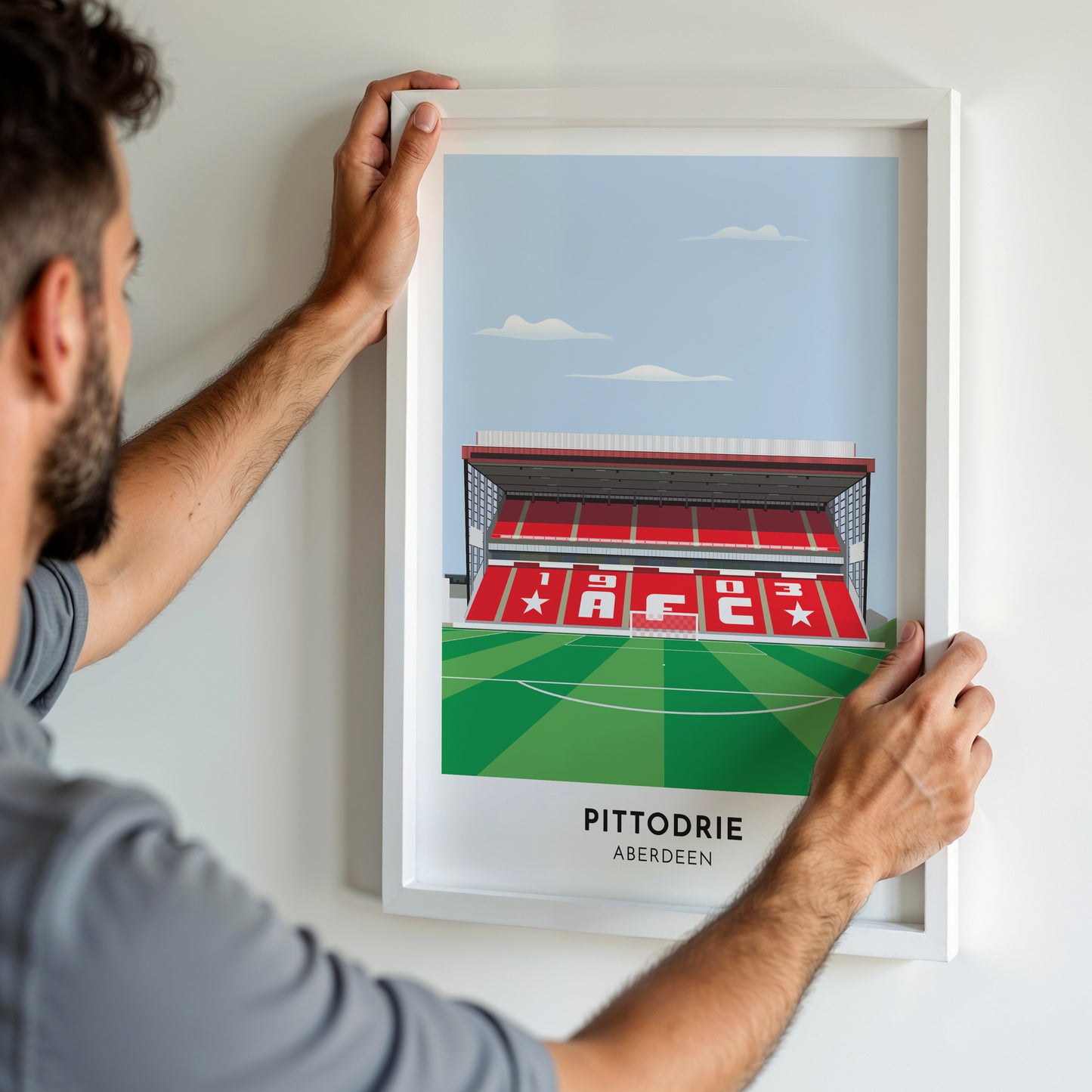 A man hanging a framed art print, featuring a contemporary style illustration of Pittodrie, home to Aberdeen.