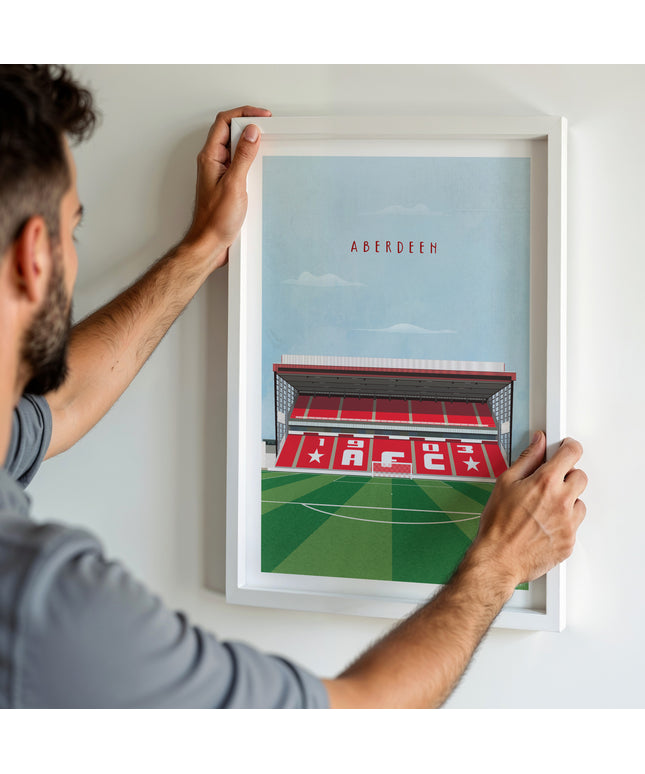A man hanging a framed art print, featuring a retro style illustration of Pittodrie, home to Aberdeen.