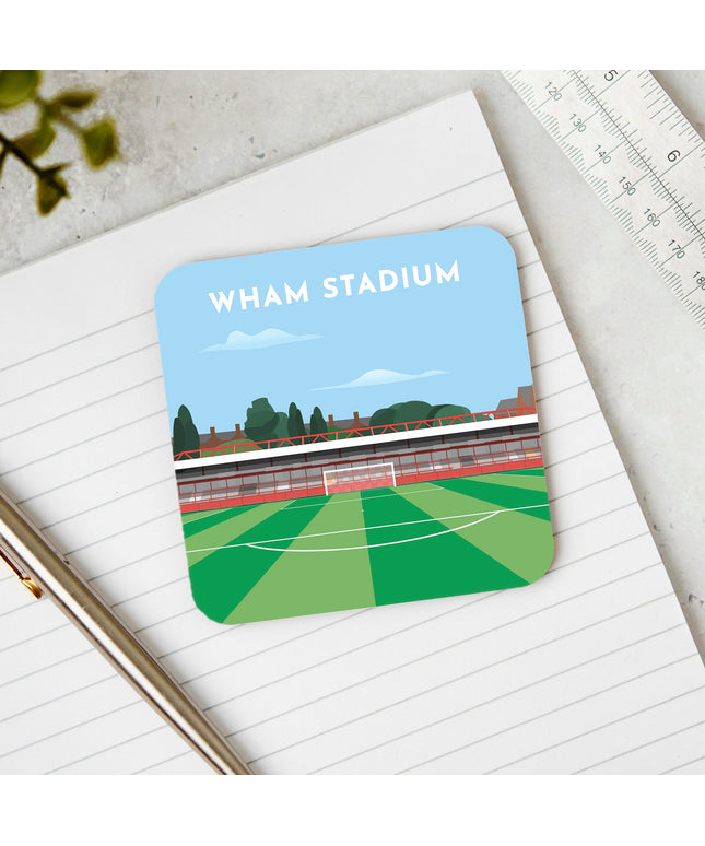 Square hardwood coaster placed on a desk, with glossy sublimation printed finish featuring contemporary illustration of Wham Stadium in Accrington, home to Accrington Stanley, showcasing a minimalist design with vibrant red stands, green football pitch, and the iconic stadium roof structure under a blue sky.