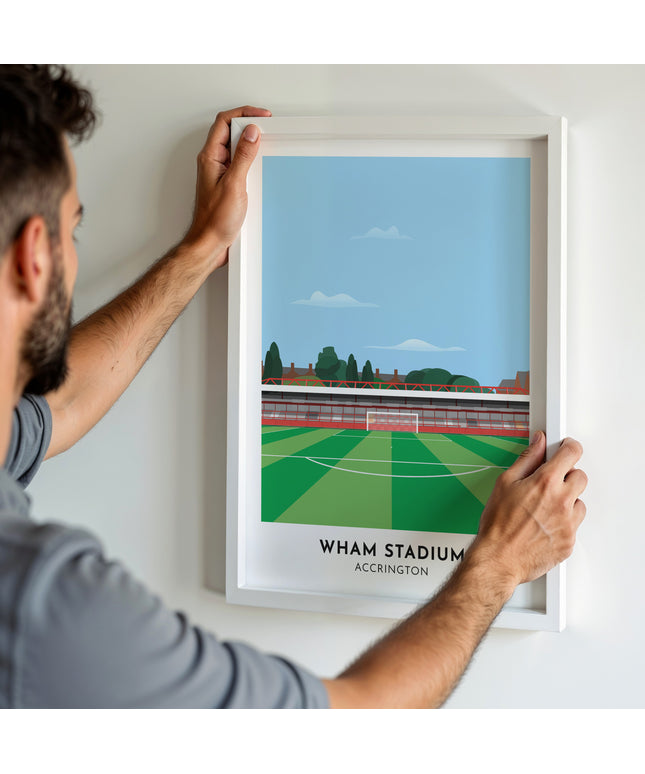 A man hanging a framed art print, featuring a contemporary style illustration of the Wham Stadium, home to Accrington Stanley