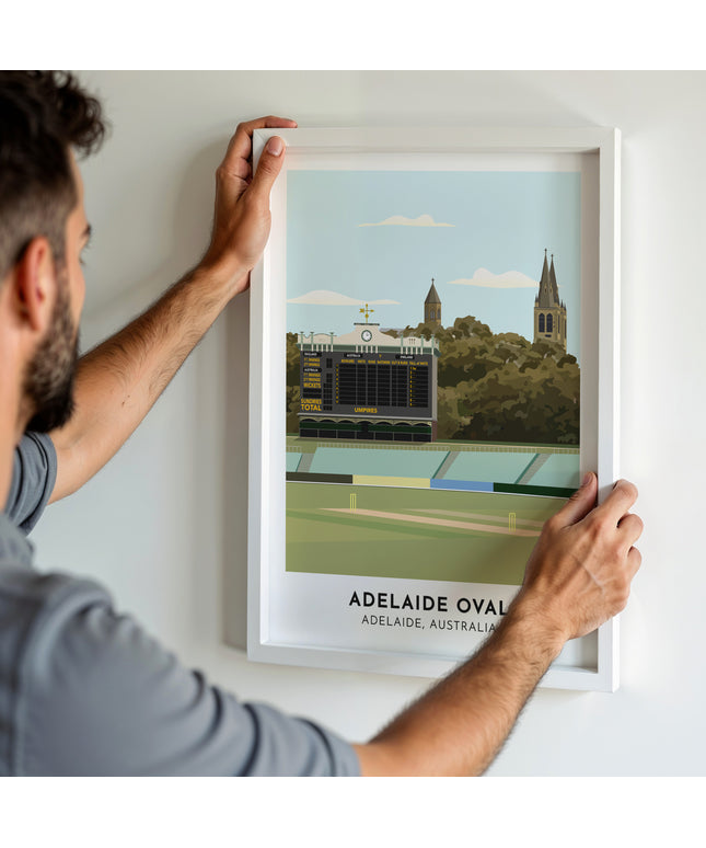 A man hanging a framed art print, featuring a contemporary style illustration of Adelaide Oval, home to South Australia.