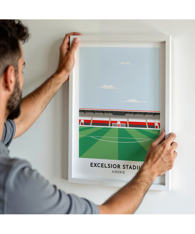 A man hanging a framed art print, featuring a contemporary style illustration of Excelsior Stadium, home to Airdrie.