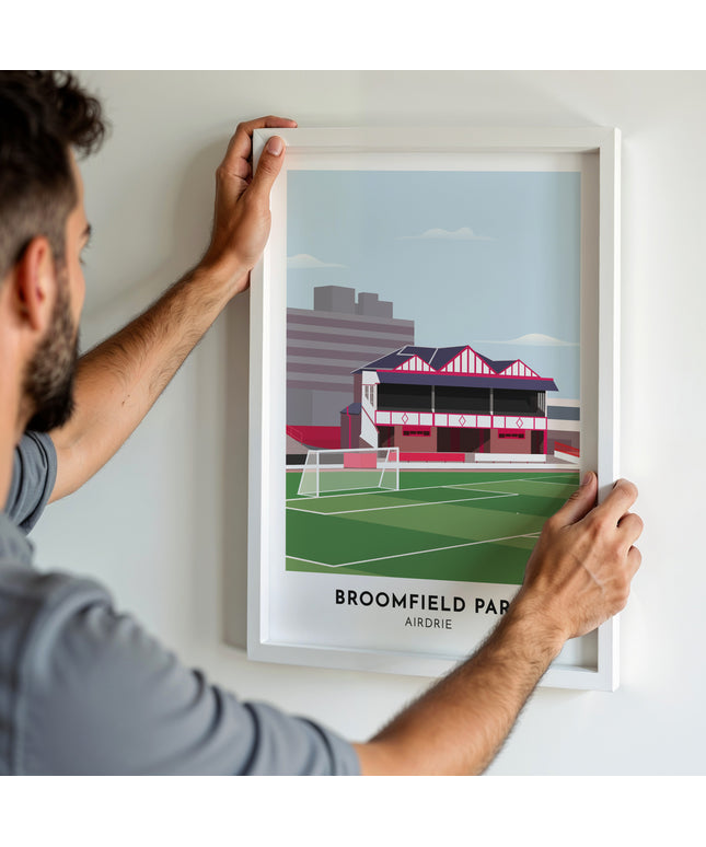 A man hanging a framed art print, featuring a contemporary style illustration of Broomfield Park, home to Airdrieonians.