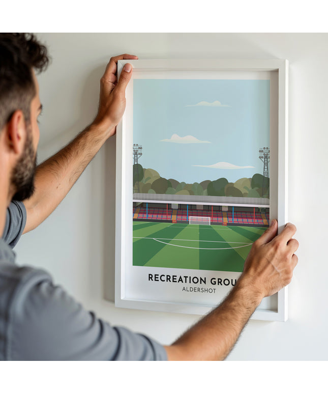 A man hanging a framed art print, featuring a contemporary style illustration of Recreation Ground, home to Aldershot Town.