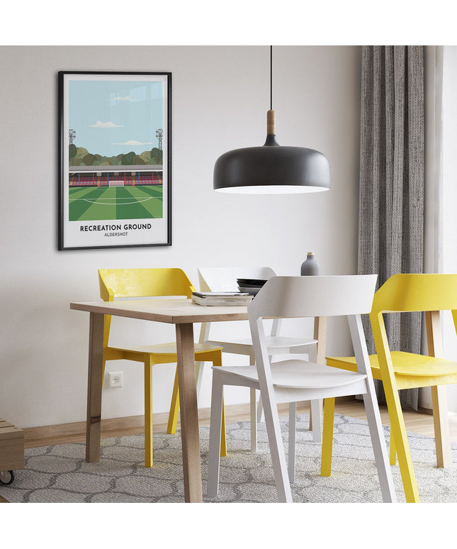Framed illustration print of Recreation Ground in Aldershot, home to Aldershot Town, showcasing a minimalist design with vibrant red and blue stands, green football pitch, under a blue sky.