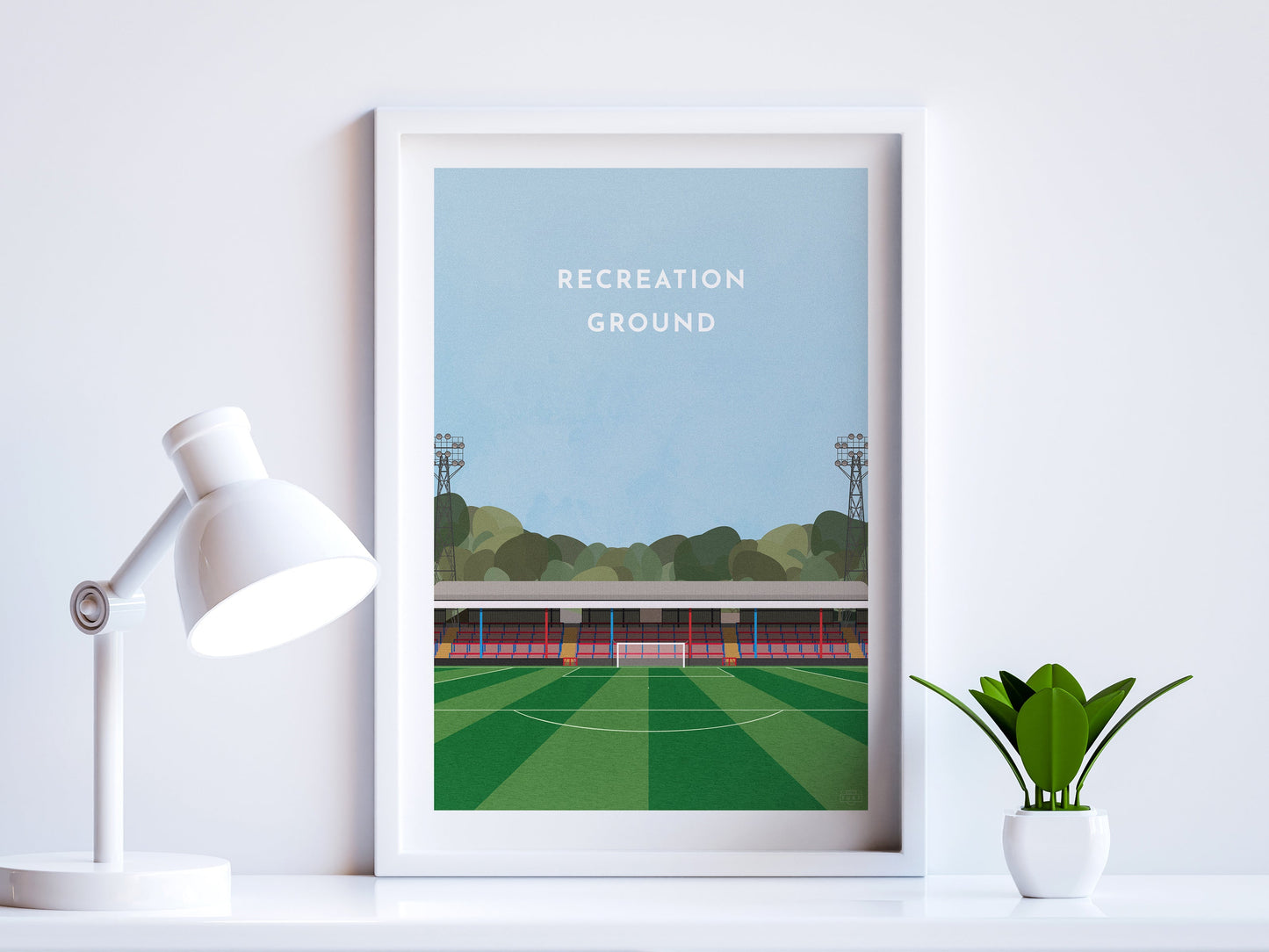 A contemporary style illustration of Aldershot Town's football stadium, the Recreation Ground, displayed as an art print within a white frame in a minimalist home office setting. 