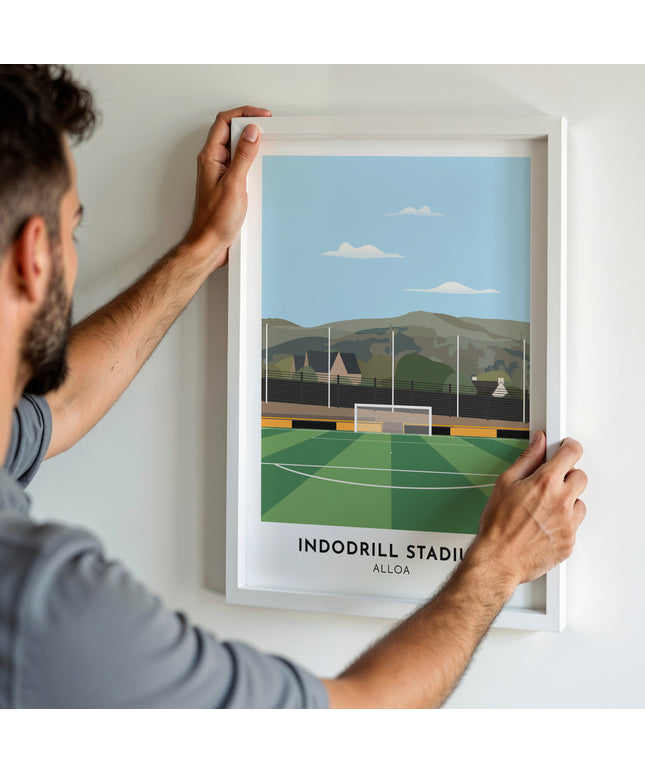 A man hanging a framed art print, featuring a contemporary style illustration of the Indodrill Stadium, home to Alloa Athletic.