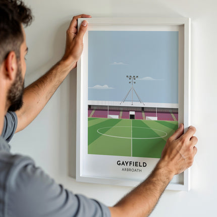 A man hanging a framed art print, featuring a contemporary style illustration of Gayfield Park, home to Arbroath.