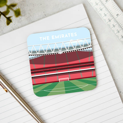 Square hardwood coaster placed on a desk, with glossy sublimation printed finish featuring contemporary illustration of The Emirates in North London, home to Arsenal, showcasing a minimalist design with vibrant red stands, green football pitch, under a blue sky.