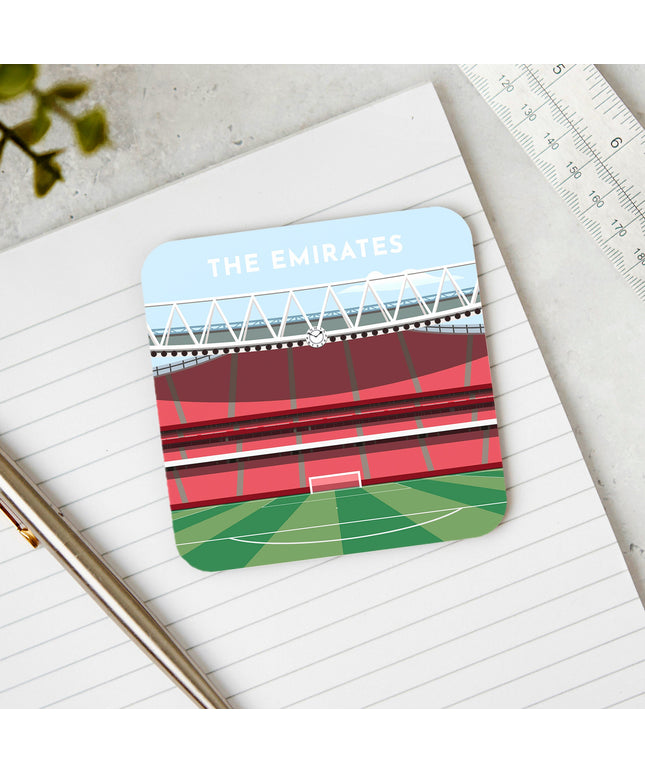 Square hardwood coaster placed on a desk, with glossy sublimation printed finish featuring contemporary illustration of The Emirates in North London, home to Arsenal, showcasing a minimalist design with vibrant red stands, green football pitch, under a blue sky.