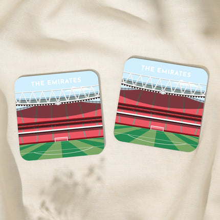 Two Square hardwood coasters laid on a tablecloth with glossy sublimation printed finish featuring contemporary illustration of The Emirates in North London, home to Arsenal, showcasing a minimalist design with vibrant red stands, green football pitch, under a blue sky.