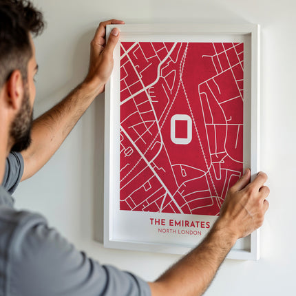 A man hanging a framed art print, featuring a contemporary style map illustration of The Emirates, home to Arsenal.
