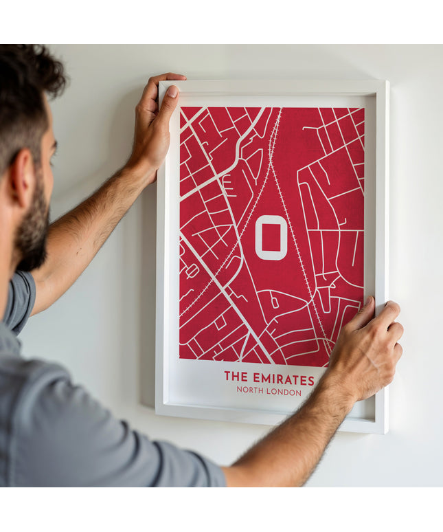 A man hanging a framed art print, featuring a contemporary style map illustration of The Emirates, home to Arsenal.