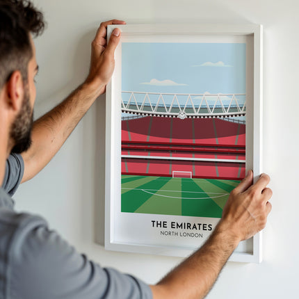 A man hanging a framed art print, featuring a contemporary style illustration of The Emirates, home to Arsenal.