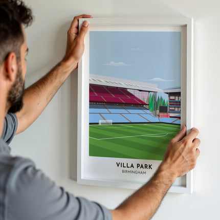 A man hanging a framed art print, featuring a contemporary style illustration of Villa Park, home to Aston V.
