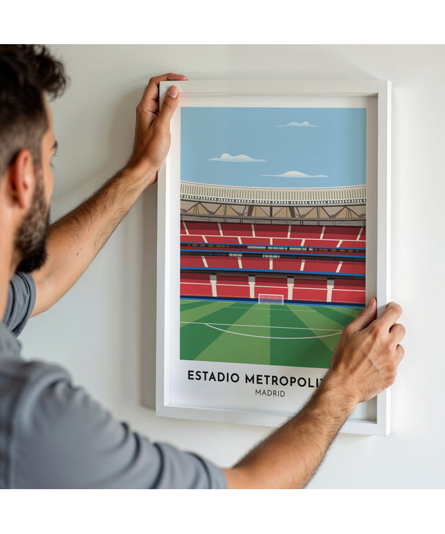 A man hanging a framed art print, featuring a contemporary style illustration of Estadio Metropolitano, home to Atlético Madrid.