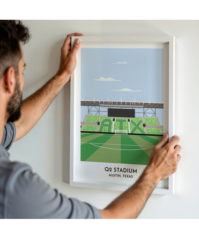 A man hanging a framed art print, featuring a contemporary style illustration of Q2 Stadium, home to Austin.
