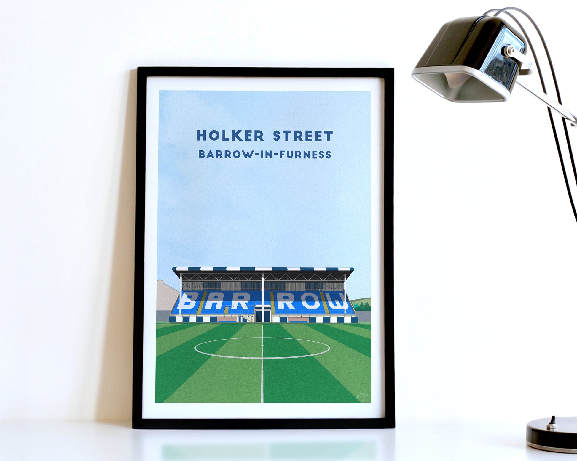 A modern style Barrow AFC Print, featuring a contemporary illustration of their home stadium the Holker Street in Barrow in Furness, displayed within a black frame.