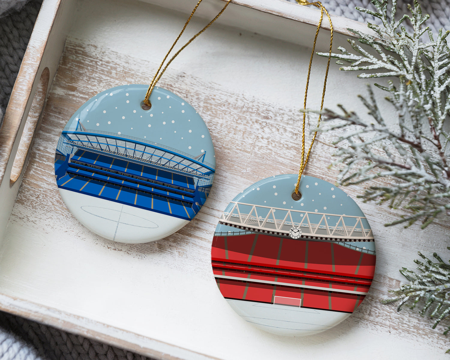 Ceramic Christmas bauble decorations featuring minimalist illustrations of football stadiums on each one, specifically Chelsea's home stadium Stamford Bridge on the left bauble, and Arsenal's Emirates Stadium on the right bauble. Caption in the image states 'request ANY stadium' referring to the product that allows you to nominate any stadium to be illustrated on the bauble in this Christmas snow scene, with snow falling in the sky and a white snow covered pitch.