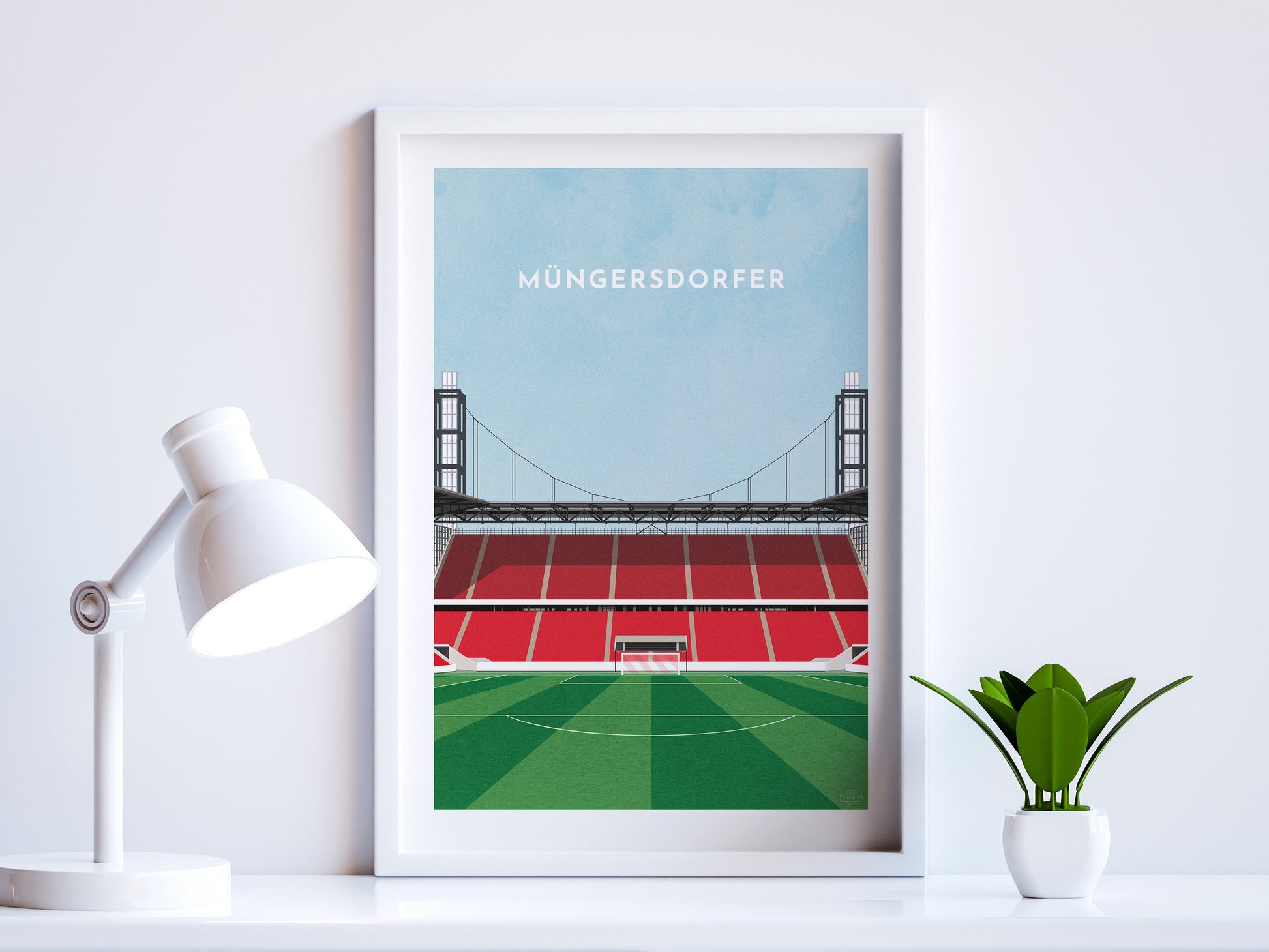 A contemporary style illustration of Cologne / Köln's football stadium, Müngersdorfer, displayed as an art print within a white frame in a minimalist home office setting. 