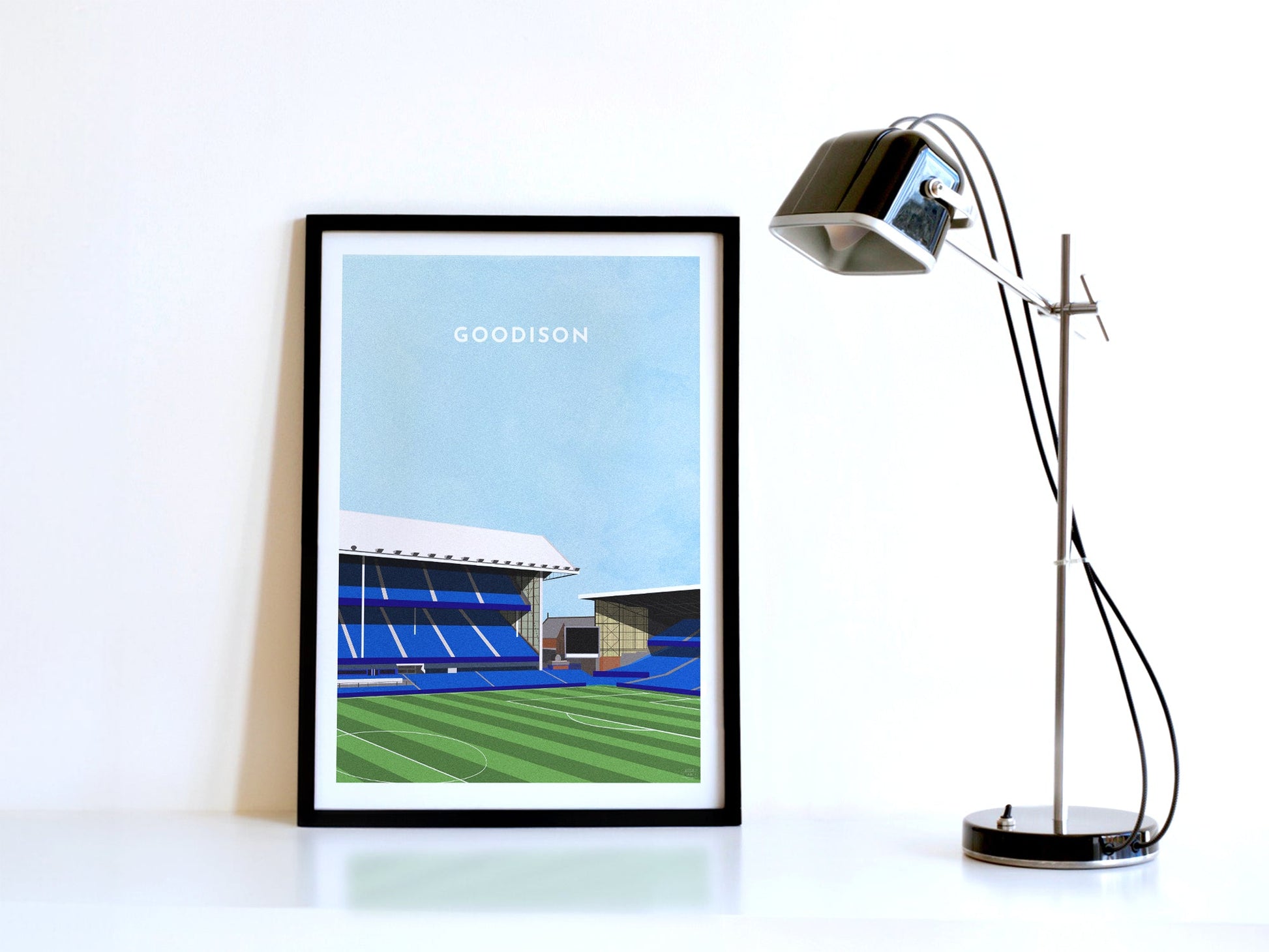 A contemporary style illustration of Everton's football stadium, Goodison Park, displayed as an art print within a black frame, next to a retro style black lamp.