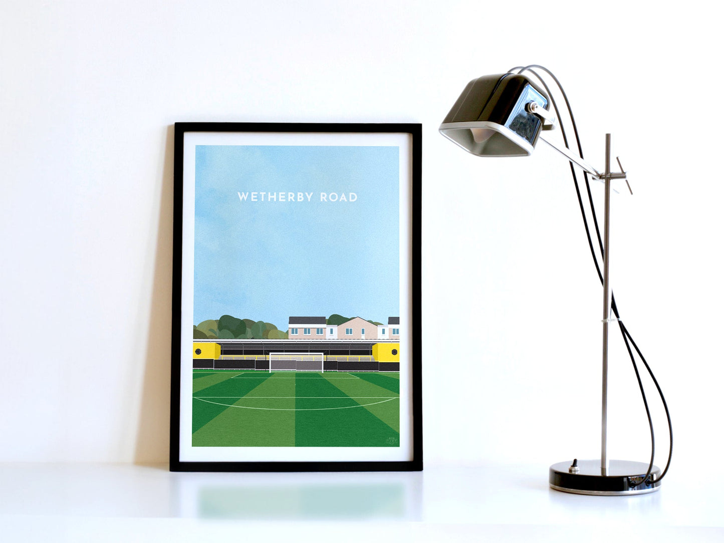 A contemporary style illustration of Harrogate Town's football ground, Wetherby Road / Exercise Stadium, displayed as an art print within a black frame, next to a retro style black lamp.