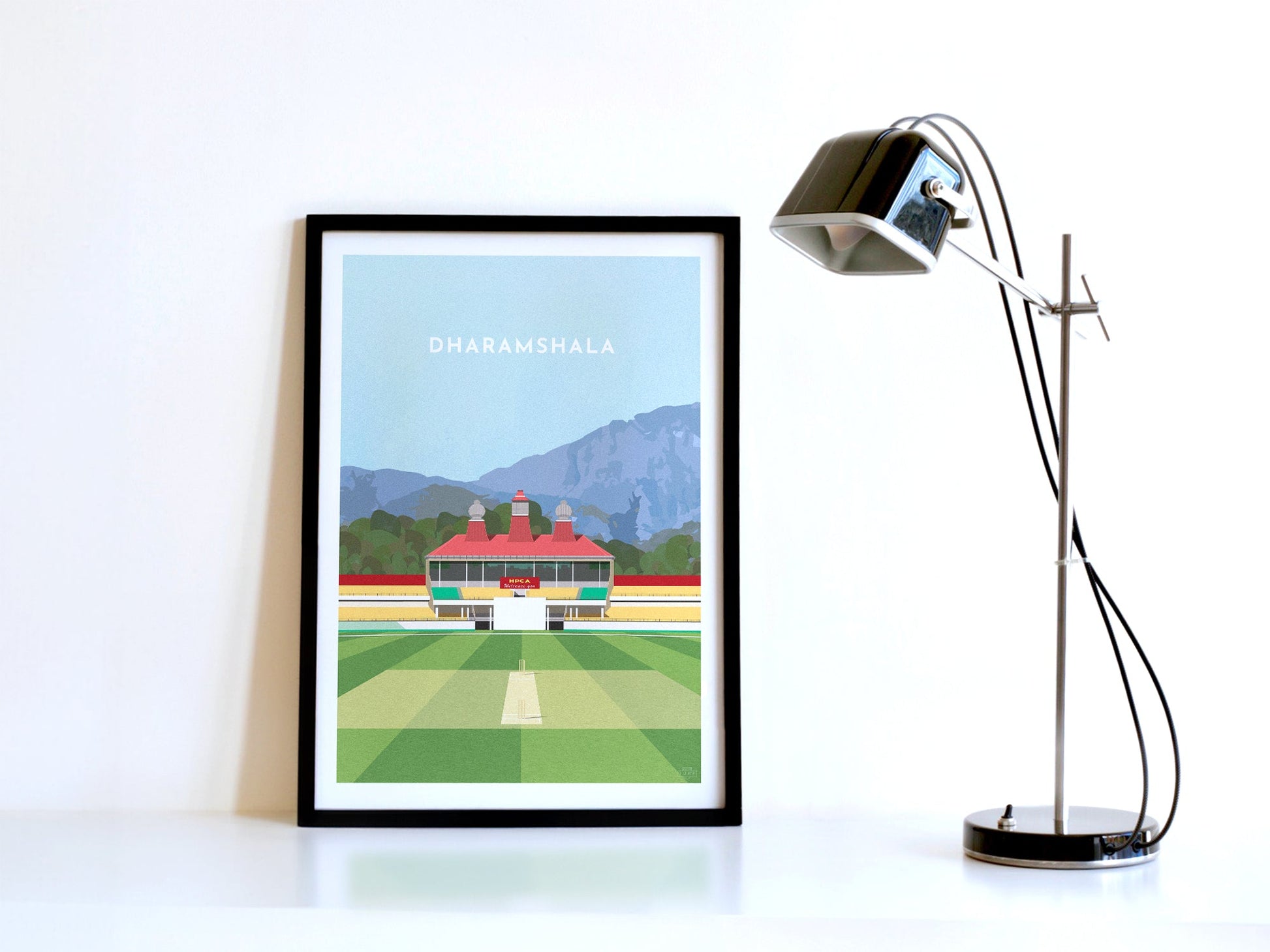 A contemporary style illustration of Himachal Pradesh's cricket stadium, the HPCA Stadium in Dharamshala, displayed as an art print within a black frame in a minimalist home office setting. 