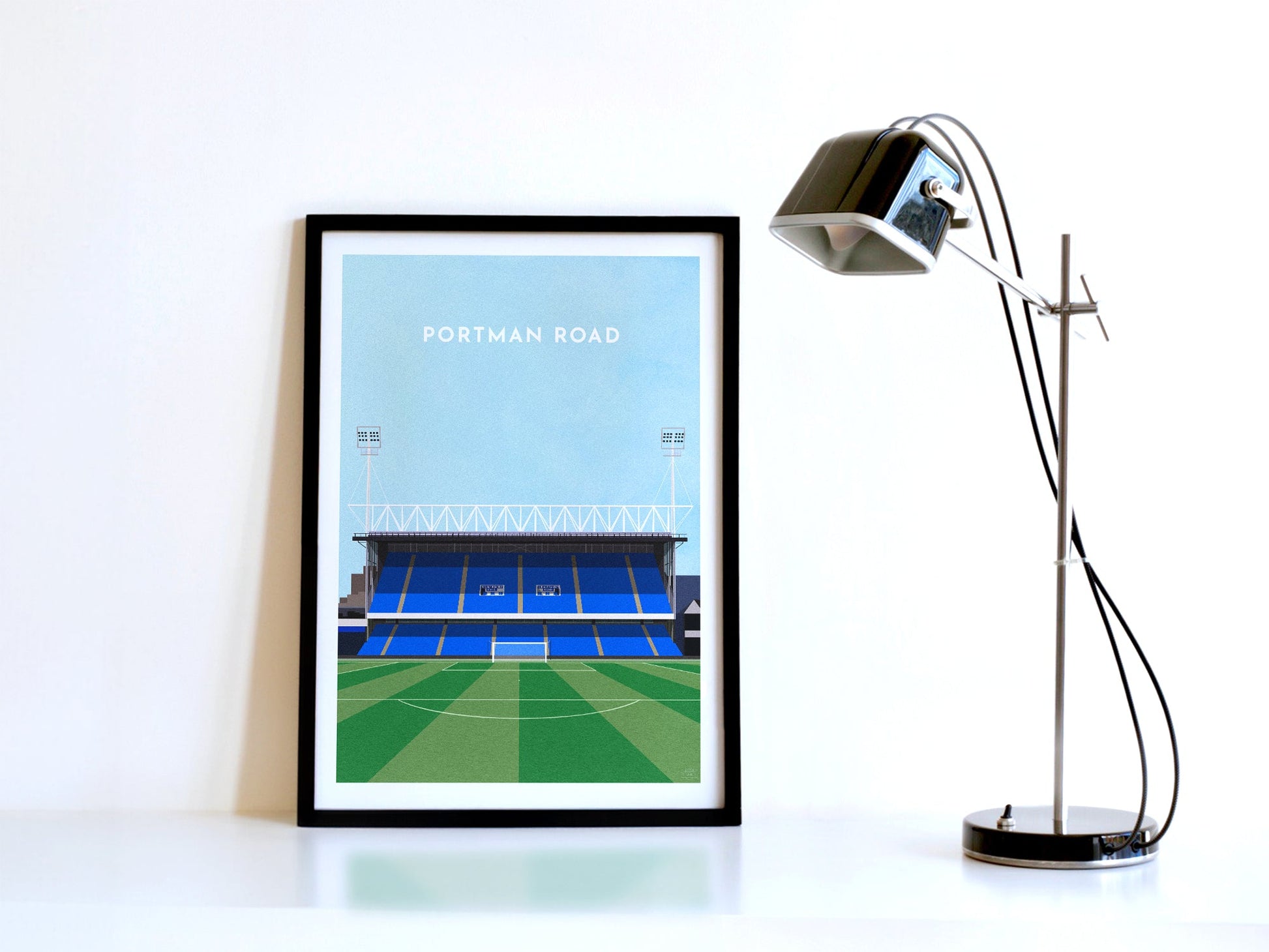 A contemporary style illustration of Ipswich's football stadium, Portman Road, displayed as an art print within a black frame, next to a retro style black lamp.