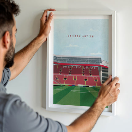 A man hanging a framed art print, featuring a retro style illustration of the Fritz-Walter Stadium, home to Kaiserslautern.