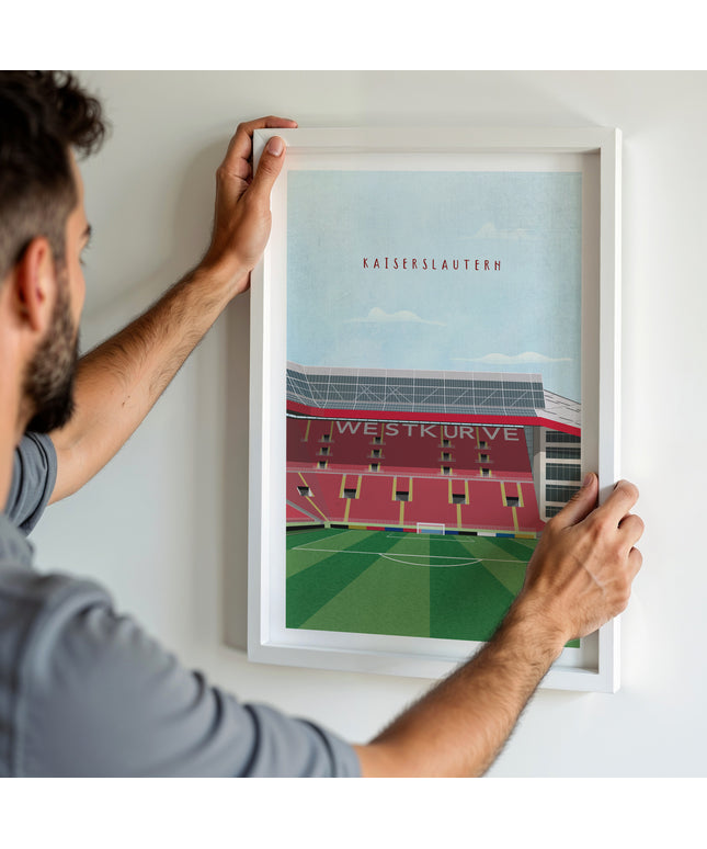 A man hanging a framed art print, featuring a retro style illustration of the Fritz-Walter Stadium, home to Kaiserslautern.