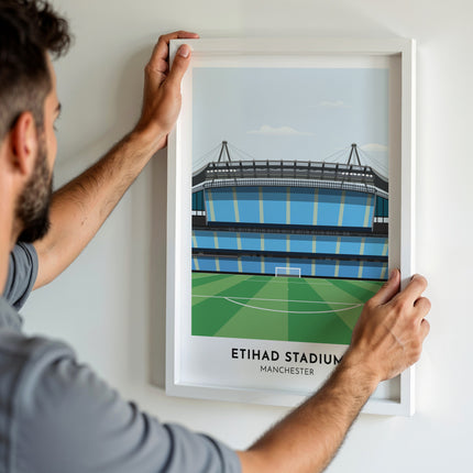 A man hanging a framed art print, featuring a contemporary style illustration of the Etihad Stadium, home to Manchester City.