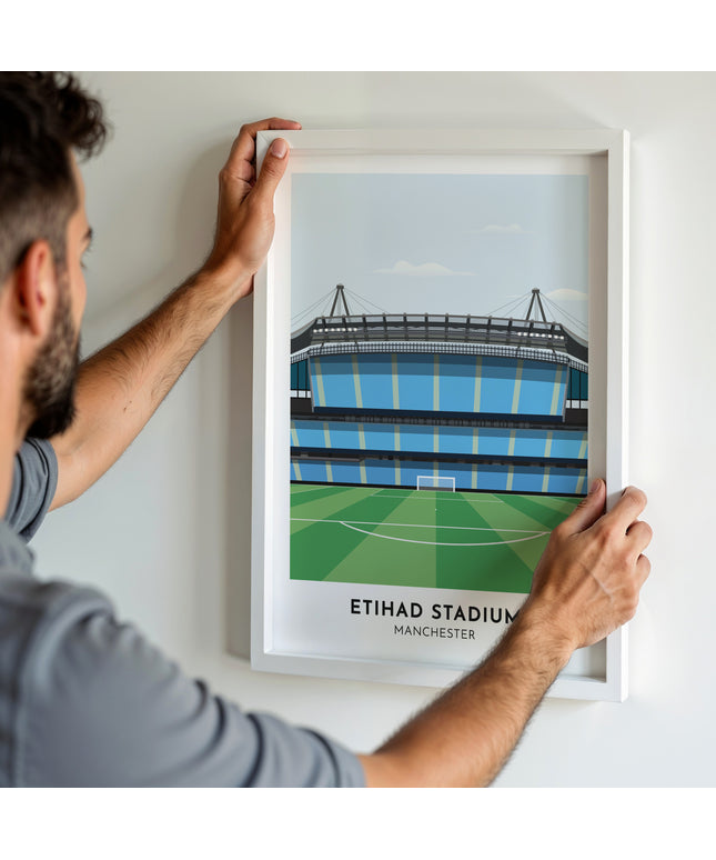 A man hanging a framed art print, featuring a contemporary style illustration of the Etihad Stadium, home to Manchester City.