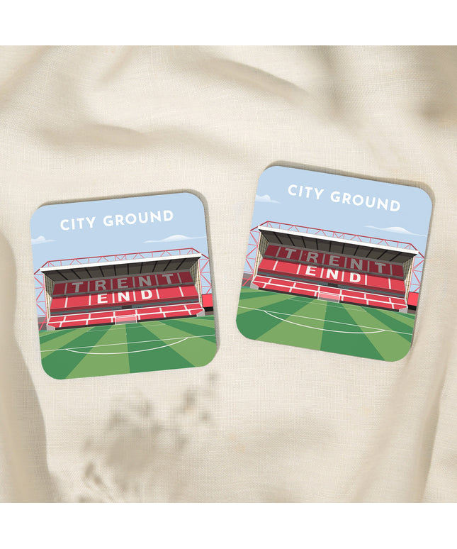 Two Square hardwood coasters laid on a tablecloth with glossy sublimation printed finish featuring contemporary illustration of City Ground in Nottingham, home to Forest, showcasing a minimalist design with vibrant COLOUR stands, green SPORT pitch, under a blue sky.